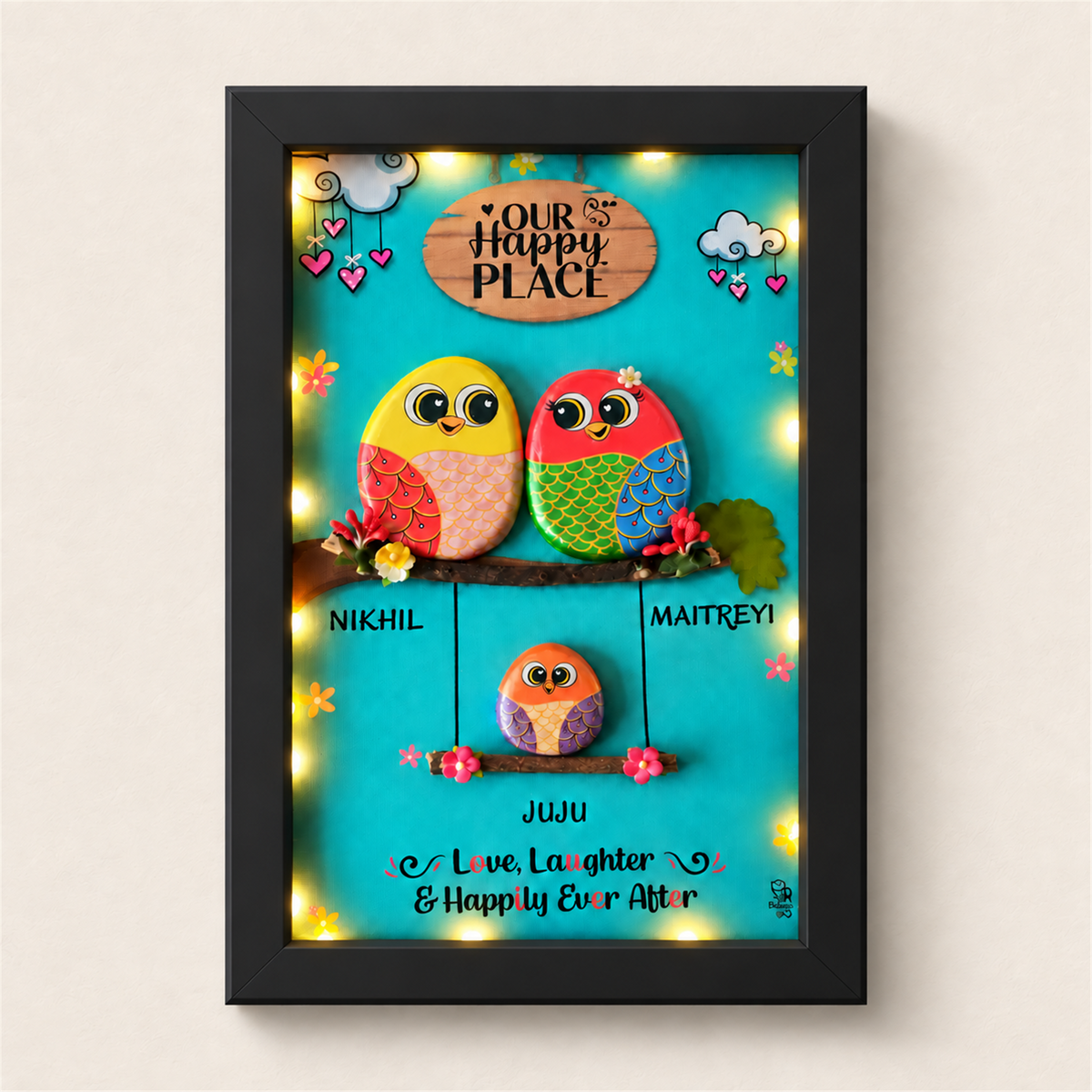 Family Joyful Nest Pebble Art: two birds above a smaller bird on a swing against a turquoise background, black frame, on a beige background