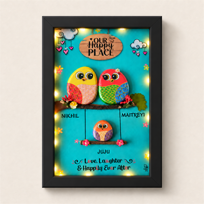 Family Joyful Nest Pebble Art: two birds above a smaller bird on a swing against a turquoise background, black frame, on a beige background