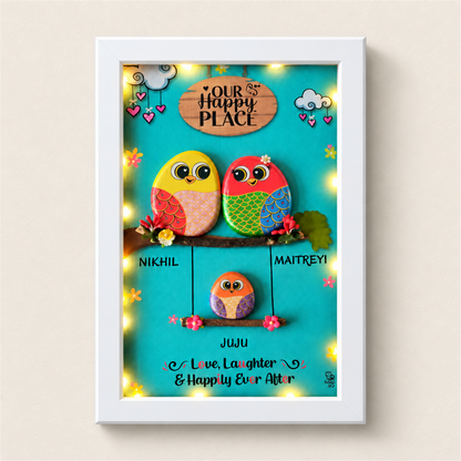 Family Joyful Nest Pebble Art: two birds above a smaller bird on a swing against a turquoise background, white frame, on a beige background