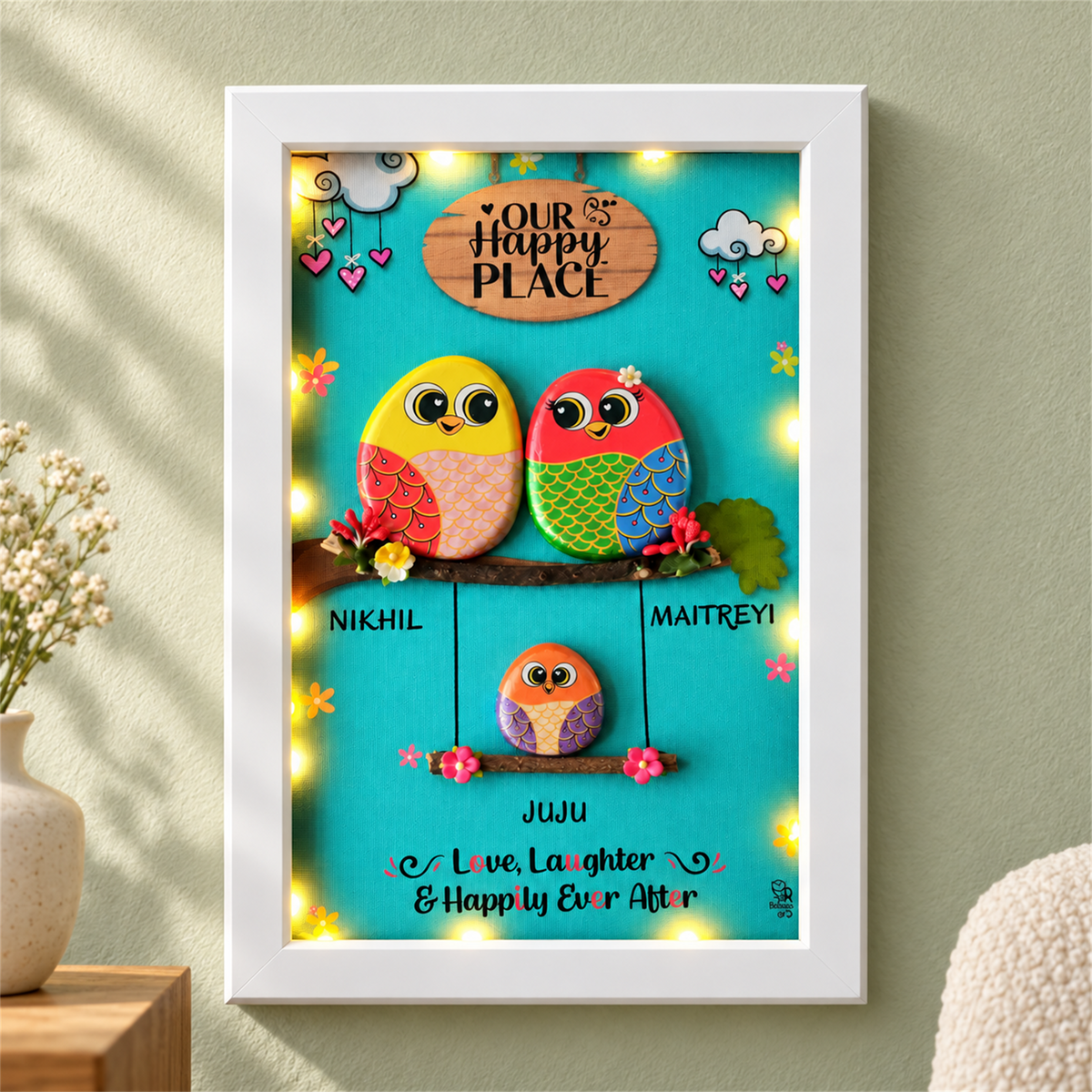 Family Joyful Nest Pebble Art: two birds above a smaller bird on a swing against a turquoise background, white frame, in a home setting